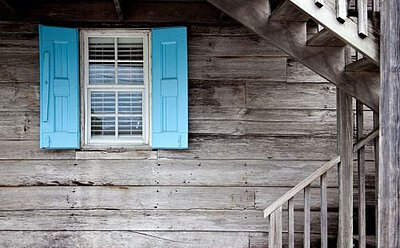 Fenster an einem Holzhaus, an welchem als Sonnenschutz sowohl Fensterläden außen als auch eine Jalousie innen sichtbar sind.